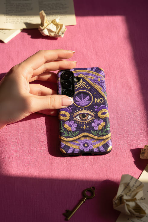A matte finish phone case featuring a 2D optical illusion of a textured, purple crochet Ouija board planchette. The design includes intricate gold faux-embroidered borders, the words YES and NO, a central all-seeing eye, and a botanical leaf motif in the center circle. Lilac purple flowers and green leaves decorate the bottom half. The case has black rubber lining around the edges and camera cutouts. It looks like a thick, handmade yarn textile but is completely flat and smooth.