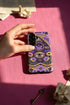 A matte finish phone case featuring a 2D optical illusion of a textured, purple crochet Ouija board planchette. The design includes intricate gold faux-embroidered borders, the words YES and NO, a central all-seeing eye, and a botanical leaf motif in the center circle. Lilac purple flowers and green leaves decorate the bottom half. The case has black rubber lining around the edges and camera cutouts. It looks like a thick, handmade yarn textile but is completely flat and smooth.