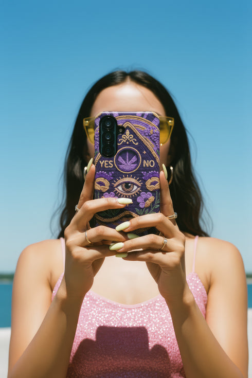 A matte finish phone case featuring a 2D optical illusion of a textured, purple crochet Ouija board planchette. The design includes intricate gold faux-embroidered borders, the words YES and NO, a central all-seeing eye, and a botanical leaf motif in the center circle. Lilac purple flowers and green leaves decorate the bottom half. The case has black rubber lining around the edges and camera cutouts. It looks like a thick, handmade yarn textile but is completely flat and smooth.