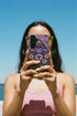 A matte finish phone case featuring a 2D optical illusion of a textured, purple crochet Ouija board planchette. The design includes intricate gold faux-embroidered borders, the words YES and NO, a central all-seeing eye, and a botanical leaf motif in the center circle. Lilac purple flowers and green leaves decorate the bottom half. The case has black rubber lining around the edges and camera cutouts. It looks like a thick, handmade yarn textile but is completely flat and smooth.
