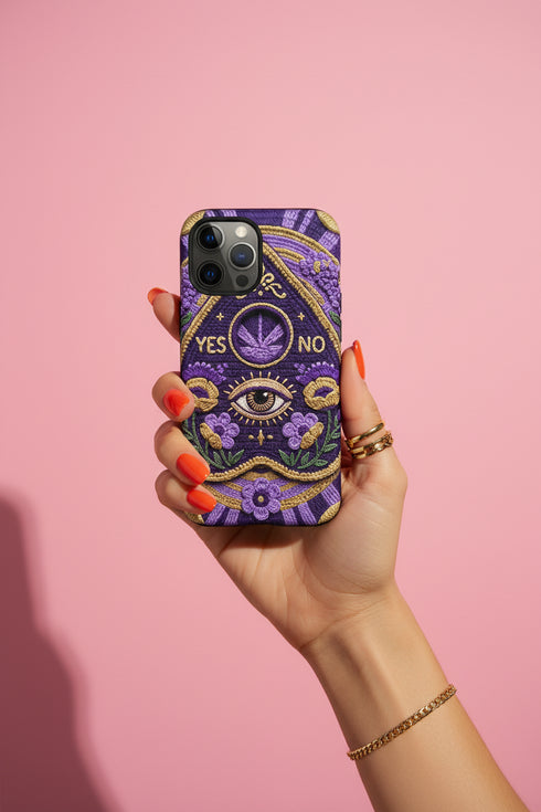 A matte finish phone case featuring a 2D optical illusion of a textured, purple crochet Ouija board planchette. The design includes intricate gold faux-embroidered borders, the words YES and NO, a central all-seeing eye, and a botanical leaf motif in the center circle. Lilac purple flowers and green leaves decorate the bottom half. The case has black rubber lining around the edges and camera cutouts. It looks like a thick, handmade yarn textile but is completely flat and smooth.