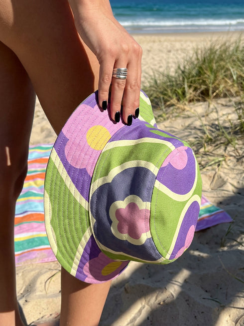Reversible bucket hat shown on a white background with two distinct prints. One side is a flowing oceanic abstract with lilac and teal wavy stripes, mint green dots, and a royal blue base. The reverse is a retro mod geometric floral in dusty rose and muted purple petals outlined in cream and olive green. The hat has a structured flat crown, a downward sloping brim with visible stitching, and a textured woven finish. Maximalist art nouveau reversible bucket hat and psychedelic festival sun hat for quirky dop
