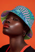 Reversible bucket hat shown on a white background with two distinct prints. One side is a flowing oceanic abstract with lilac and teal wavy stripes, mint green dots, and a royal blue base. The reverse is a retro mod geometric floral in dusty rose and muted purple petals outlined in cream and olive green. The hat has a structured flat crown, a downward sloping brim with visible stitching, and a textured woven finish. Maximalist art nouveau reversible bucket hat and psychedelic festival sun hat for quirky dop