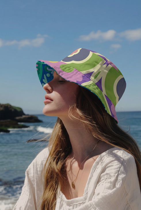 Reversible bucket hat shown on a white background with two distinct prints. One side is a flowing oceanic abstract with lilac and teal wavy stripes, mint green dots, and a royal blue base. The reverse is a retro mod geometric floral in dusty rose and muted purple petals outlined in cream and olive green. The hat has a structured flat crown, a downward sloping brim with visible stitching, and a textured woven finish. Maximalist art nouveau reversible bucket hat and psychedelic festival sun hat for quirky dop