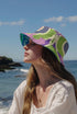 Reversible bucket hat shown on a white background with two distinct prints. One side is a flowing oceanic abstract with lilac and teal wavy stripes, mint green dots, and a royal blue base. The reverse is a retro mod geometric floral in dusty rose and muted purple petals outlined in cream and olive green. The hat has a structured flat crown, a downward sloping brim with visible stitching, and a textured woven finish. Maximalist art nouveau reversible bucket hat and psychedelic festival sun hat for quirky dop