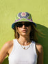 Reversible bucket hat shown on a white background with two distinct prints. One side is a flowing oceanic abstract with lilac and teal wavy stripes, mint green dots, and a royal blue base. The reverse is a retro mod geometric floral in dusty rose and muted purple petals outlined in cream and olive green. The hat has a structured flat crown, a downward sloping brim with visible stitching, and a textured woven finish. Maximalist art nouveau reversible bucket hat and psychedelic festival sun hat for quirky dop
