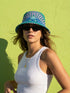 Reversible bucket hat shown on a white background with two distinct prints. One side is a flowing oceanic abstract with lilac and teal wavy stripes, mint green dots, and a royal blue base. The reverse is a retro mod geometric floral in dusty rose and muted purple petals outlined in cream and olive green. The hat has a structured flat crown, a downward sloping brim with visible stitching, and a textured woven finish. Maximalist art nouveau reversible bucket hat and psychedelic festival sun hat for quirky dop