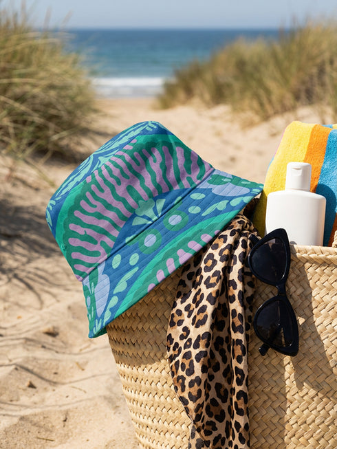 Reversible bucket hat shown on a white background with two distinct prints. One side is a flowing oceanic abstract with lilac and teal wavy stripes, mint green dots, and a royal blue base. The reverse is a retro mod geometric floral in dusty rose and muted purple petals outlined in cream and olive green. The hat has a structured flat crown, a downward sloping brim with visible stitching, and a textured woven finish. Maximalist art nouveau reversible bucket hat and psychedelic festival sun hat for quirky dop