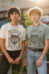 Vintage style graphic t-shirt with a large snarling wolf face printed in dark charcoal ink. A curved banner above reads RAISED BY WOLVES and a banner below reads RADICALIZED BY LEGISLATION, framed by stylized flames and scattered stars. Classic crewneck short sleeve cotton tee with a relaxed fit and seamless sides, available in soft pink, pale green, and ivory. Feminist protest apparel and activist statement tee for reproductive rights, marches, and everyday quiet rage.