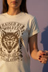 Vintage style graphic t-shirt with a large snarling wolf face printed in dark charcoal ink. A curved banner above reads RAISED BY WOLVES and a banner below reads RADICALIZED BY LEGISLATION, framed by stylized flames and scattered stars. Classic crewneck short sleeve cotton tee with a relaxed fit and seamless sides, available in soft pink, pale green, and ivory. Feminist protest apparel and activist statement tee for reproductive rights, marches, and everyday quiet rage.