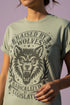 Vintage style graphic t-shirt with a large snarling wolf face printed in dark charcoal ink. A curved banner above reads RAISED BY WOLVES and a banner below reads RADICALIZED BY LEGISLATION, framed by stylized flames and scattered stars. Classic crewneck short sleeve cotton tee with a relaxed fit and seamless sides, available in soft pink, pale green, and ivory. Feminist protest apparel and activist statement tee for reproductive rights, marches, and everyday quiet rage.