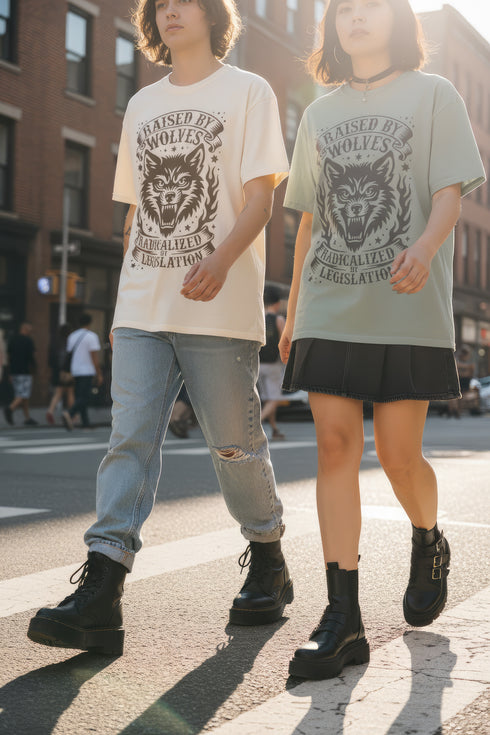 Vintage style graphic t-shirt with a large snarling wolf face printed in dark charcoal ink. A curved banner above reads RAISED BY WOLVES and a banner below reads RADICALIZED BY LEGISLATION, framed by stylized flames and scattered stars. Classic crewneck short sleeve cotton tee with a relaxed fit and seamless sides, available in soft pink, pale green, and ivory. Feminist protest apparel and activist statement tee for reproductive rights, marches, and everyday quiet rage.