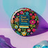Round pinback button featuring a teal book with "Read Banned Books" in gold lettering on black background, surrounded by vibrant embroidery-style flowers in pink, orange, and blue. Literary protest pin with glossy finish and safety pin backing, available in 2" and 3" sizes for bookish fashion statements.