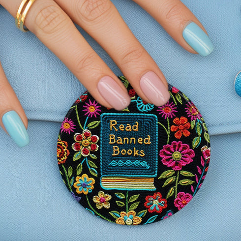 Round pinback button featuring a teal book with "Read Banned Books" in gold lettering on black background, surrounded by vibrant embroidery-style flowers in pink, orange, and blue. Literary protest pin with glossy finish and safety pin backing, available in 2" and 3" sizes for bookish fashion statements.