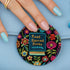 Round pinback button featuring a teal book with "Read Banned Books" in gold lettering on black background, surrounded by vibrant embroidery-style flowers in pink, orange, and blue. Literary protest pin with glossy finish and safety pin backing, available in 2" and 3" sizes for bookish fashion statements.