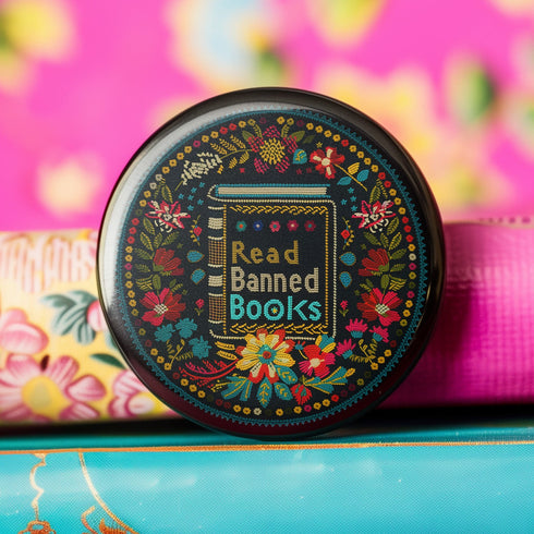 Round black pinback button with vibrant Mexican folk art-inspired embroidery design featuring red, yellow, blue and teal flowers surrounding a gold book spine with "Read Banned Books" text. Literary protest accessory with glossy finish and safety pin backing, available in 2" and 3" sizes.