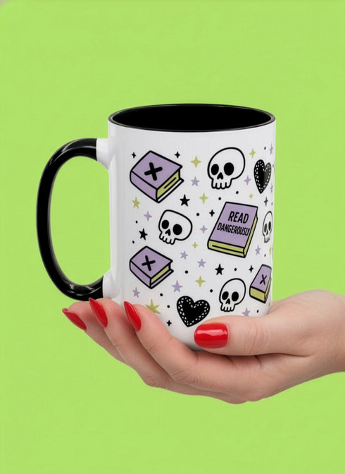 A white ceramic mug with a black interior and black C-shaped handle. The mug features a full wrap-around design in a pastel goth style. The repeating pattern includes cartoon illustrations of white skulls, black stitched hearts, and pastel purple books with an 'X' on the cover. One book is open and displays the words 'READ DANGEROUSLY'. The graphics are scattered with small pastel purple and pale yellow-green stars, creating a spooky and cute aesthetic.