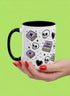 A white ceramic mug with a black interior and black C-shaped handle. The mug features a full wrap-around design in a pastel goth style. The repeating pattern includes cartoon illustrations of white skulls, black stitched hearts, and pastel purple books with an 'X' on the cover. One book is open and displays the words 'READ DANGEROUSLY'. The graphics are scattered with small pastel purple and pale yellow-green stars, creating a spooky and cute aesthetic.