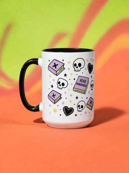 A white ceramic mug with a black interior and black C-shaped handle. The mug features a full wrap-around design in a pastel goth style. The repeating pattern includes cartoon illustrations of white skulls, black stitched hearts, and pastel purple books with an 'X' on the cover. One book is open and displays the words 'READ DANGEROUSLY'. The graphics are scattered with small pastel purple and pale yellow-green stars, creating a spooky and cute aesthetic.