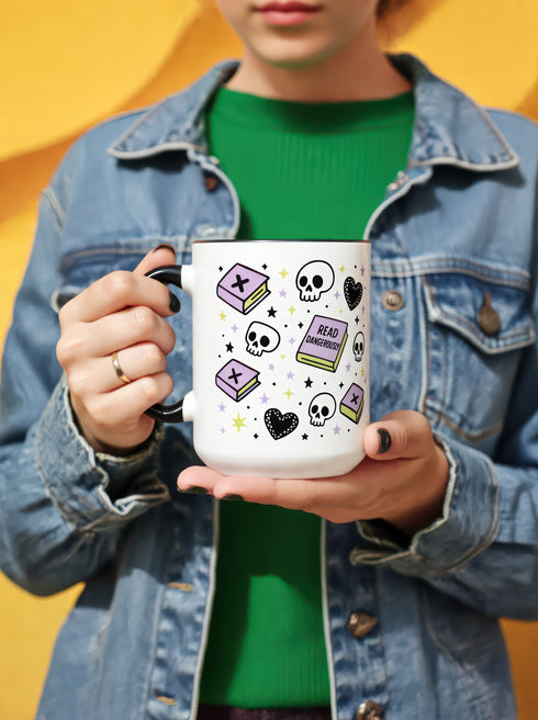 A white ceramic mug with a black interior and black C-shaped handle. The mug features a full wrap-around design in a pastel goth style. The repeating pattern includes cartoon illustrations of white skulls, black stitched hearts, and pastel purple books with an 'X' on the cover. One book is open and displays the words 'READ DANGEROUSLY'. The graphics are scattered with small pastel purple and pale yellow-green stars, creating a spooky and cute aesthetic.