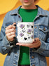 A white ceramic mug with a black interior and black C-shaped handle. The mug features a full wrap-around design in a pastel goth style. The repeating pattern includes cartoon illustrations of white skulls, black stitched hearts, and pastel purple books with an 'X' on the cover. One book is open and displays the words 'READ DANGEROUSLY'. The graphics are scattered with small pastel purple and pale yellow-green stars, creating a spooky and cute aesthetic.