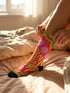 A pair of vibrant crew socks featuring a maximalist 1970s inspired collage pattern. The design includes blocks of hot pink with white daisy flowers and yellow centers, clashing playfully with panels of mustard yellow and orange tiger stripes. Wavy psychedelic lines in grass green and orange frame the top cuff. The socks have a solid black heel and toe cap. The fabric is a smooth, stretchy polyester blend with a flat knit texture and bright colors.