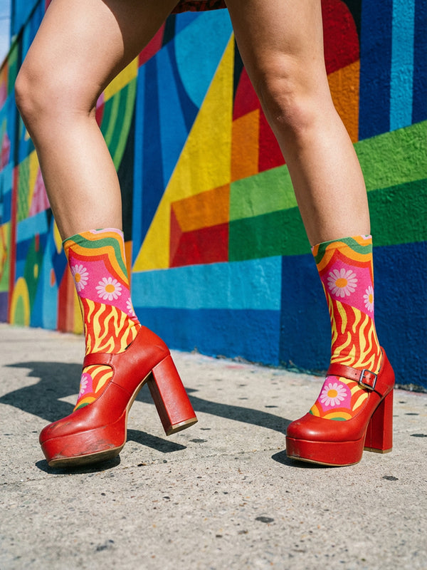 A pair of vibrant crew socks featuring a maximalist 1970s inspired collage pattern. The design includes blocks of hot pink with white daisy flowers and yellow centers, clashing playfully with panels of mustard yellow and orange tiger stripes. Wavy psychedelic lines in grass green and orange frame the top cuff. The socks have a solid black heel and toe cap. The fabric is a smooth, stretchy polyester blend with a flat knit texture and bright colors.