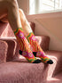 A pair of vibrant crew socks featuring a maximalist 1970s inspired collage pattern. The design includes blocks of hot pink with white daisy flowers and yellow centers, clashing playfully with panels of mustard yellow and orange tiger stripes. Wavy psychedelic lines in grass green and orange frame the top cuff. The socks have a solid black heel and toe cap. The fabric is a smooth, stretchy polyester blend with a flat knit texture and bright colors.