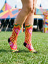A pair of vibrant crew socks featuring a maximalist 1970s inspired collage pattern. The design includes blocks of hot pink with white daisy flowers and yellow centers, clashing playfully with panels of mustard yellow and orange tiger stripes. Wavy psychedelic lines in grass green and orange frame the top cuff. The socks have a solid black heel and toe cap. The fabric is a smooth, stretchy polyester blend with a flat knit texture and bright colors.