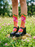 A pair of vibrant crew socks featuring a maximalist 1970s inspired collage pattern. The design includes blocks of hot pink with white daisy flowers and yellow centers, clashing playfully with panels of mustard yellow and orange tiger stripes. Wavy psychedelic lines in grass green and orange frame the top cuff. The socks have a solid black heel and toe cap. The fabric is a smooth, stretchy polyester blend with a flat knit texture and bright colors.
