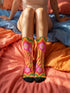 A pair of vibrant crew socks featuring a maximalist 1970s inspired collage pattern. The design includes blocks of hot pink with white daisy flowers and yellow centers, clashing playfully with panels of mustard yellow and orange tiger stripes. Wavy psychedelic lines in grass green and orange frame the top cuff. The socks have a solid black heel and toe cap. The fabric is a smooth, stretchy polyester blend with a flat knit texture and bright colors.