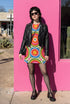 Short sleeve crew neck t-shirt dress on a cream background with an allover retro mod flower medallion print. Bold 60s daisies in red, yellow, and white sit at the center of concentric rainbow rings in lime green, hot pink, orange, and royal blue, with small magenta squares and tiny red checkerboard accents between the motifs. Above-the-knee straight fit silhouette in a 95 percent cotton, 5 percent elastane heavyweight stretch blend. Groovy retro flower power dress for dopamine dressing and easy summer outfi