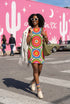 Short sleeve crew neck t-shirt dress on a cream background with an allover retro mod flower medallion print. Bold 60s daisies in red, yellow, and white sit at the center of concentric rainbow rings in lime green, hot pink, orange, and royal blue, with small magenta squares and tiny red checkerboard accents between the motifs. Above-the-knee straight fit silhouette in a 95 percent cotton, 5 percent elastane heavyweight stretch blend. Groovy retro flower power dress for dopamine dressing and easy summer outfi