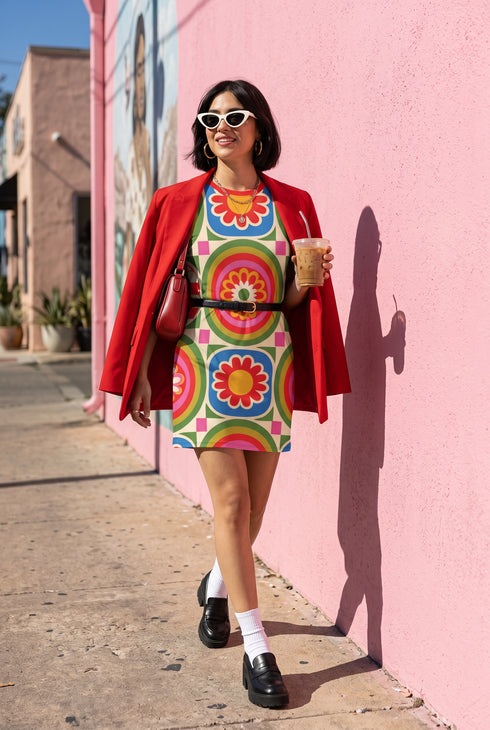 Short sleeve crew neck t-shirt dress on a cream background with an allover retro mod flower medallion print. Bold 60s daisies in red, yellow, and white sit at the center of concentric rainbow rings in lime green, hot pink, orange, and royal blue, with small magenta squares and tiny red checkerboard accents between the motifs. Above-the-knee straight fit silhouette in a 95 percent cotton, 5 percent elastane heavyweight stretch blend. Groovy retro flower power dress for dopamine dressing and easy summer outfi