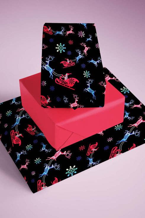 Christmas wrapping paper with a black background featuring colorful, stylized reindeer and Santa's sleigh designs. The reindeer and sleigh silhouettes are filled with skeletal patterns in shades of blue, pink, and red. Multicolored snowflakes in various sizes are scattered throughout. The paper has a whimsical, slightly gothic aesthetic that blends traditional holiday imagery with a unique, edgy twist.