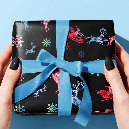Christmas wrapping paper with a black background featuring colorful, stylized reindeer and Santa's sleigh designs. The reindeer and sleigh silhouettes are filled with skeletal patterns in shades of blue, pink, and red. Multicolored snowflakes in various sizes are scattered throughout. The paper has a whimsical, slightly gothic aesthetic that blends traditional holiday imagery with a unique, edgy twist.