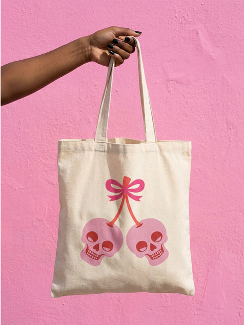Black 15 x 16 inch 100% cotton tote bag with a kawaii pastel goth cherry-skull design printed on the front. The artwork features two soft pink skulls shaped like cherries, connected by red stems and topped with a bright pink bow. Cute, spooky, coquette, and alt-inspired everyday tote.