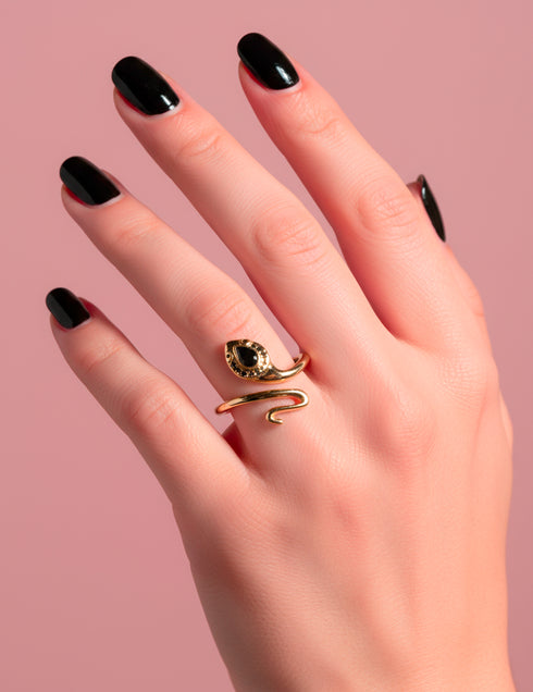 Close-up of a gold-plated Cobra Head Wrap Ring featuring a black teardrop-shaped zircon gemstone, wrapped in a coiled snake design. The ring rests on a hand with black nails against a pink background. Artistic serpent jewelry by Honey Dazed with a bold, modern gothic vibe.