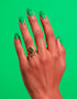 A gold-plated serpent ring with a shimmering green zircon gemstone shaped like a cobra’s head, worn on a hand with dark nails. The adjustable wrap design and luxe detailing are shown against a pink backdrop. Witchy statement jewelry by Honey Dazed.