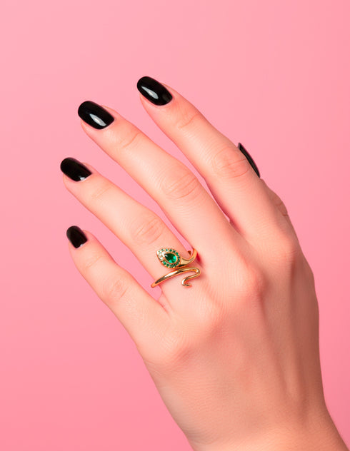 A gold-plated serpent ring with a shimmering green zircon gemstone shaped like a cobra’s head, worn on a hand with dark nails. The adjustable wrap design and luxe detailing are shown against a pink backdrop. Witchy statement jewelry by Honey Dazed.