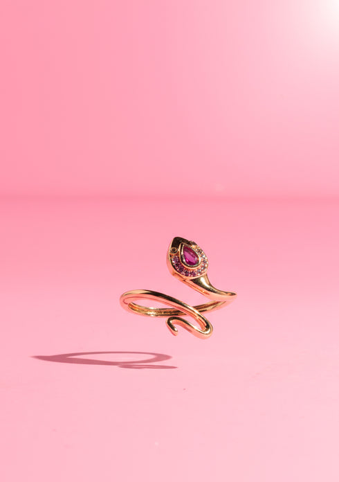 A pink zircon Cobra Head Wrap Ring in gold-plated copper, coiled elegantly around a model’s finger. The teardrop gemstone and intricate snake detailing shine against a blush-pink background. Perfect for alt fashion and bold statement looks.