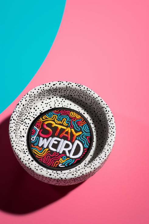 A round embroidered patch with a black background featuring the phrase “STAY WEIRD” in bold, multicolored lettering. "STAY" is stitched in a gradient of red, orange, and yellow, while "WEIRD" is in white. Surrounding the text are wavy, neon-like lines in blue, pink, yellow, and orange, creating a vibrant, psychedelic effect. The patch has a thick black border and is designed for iron-on or sew-on application, making it perfect for jackets, backpacks, denim, and accessories.