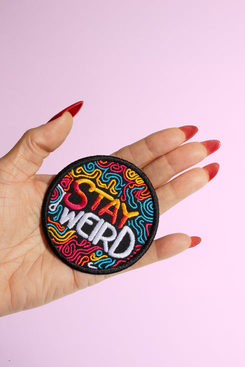 A round embroidered patch with a black background featuring the phrase “STAY WEIRD” in bold, multicolored lettering. "STAY" is stitched in a gradient of red, orange, and yellow, while "WEIRD" is in white. Surrounding the text are wavy, neon-like lines in blue, pink, yellow, and orange, creating a vibrant, psychedelic effect. The patch has a thick black border and is designed for iron-on or sew-on application, making it perfect for jackets, backpacks, denim, and accessories.