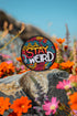 A round embroidered patch with a black background featuring the phrase “STAY WEIRD” in bold, multicolored lettering. "STAY" is stitched in a gradient of red, orange, and yellow, while "WEIRD" is in white. Surrounding the text are wavy, neon-like lines in blue, pink, yellow, and orange, creating a vibrant, psychedelic effect. The patch has a thick black border and is designed for iron-on or sew-on application, making it perfect for jackets, backpacks, denim, and accessories.