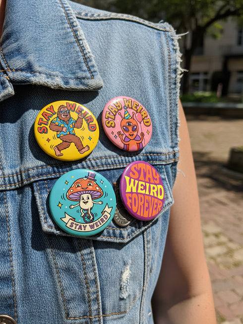 Set of 4 bright retro pinback buttons 2.25 inch. 1: Yellow button with cartoon Bigfoot in blue Hawaiian shirt and sunglasses striding under "Stay Weird" text. 2: Pink button with orange three-eyed alien in party hat holding a balloon. 3: Teal button with smiling mushroom in top hat riding a unicycle with "Stay Weird" banner. 4: Purple button with orange and yellow psychedelic 70s font reading "Stay Weird Forever." High gloss finish metal pins for backpacks, jackets, and weirdcore gifts.