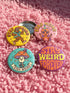 Set of 4 bright retro pinback buttons 2.25 inch. 1: Yellow button with cartoon Bigfoot in blue Hawaiian shirt and sunglasses striding under "Stay Weird" text. 2: Pink button with orange three-eyed alien in party hat holding a balloon. 3: Teal button with smiling mushroom in top hat riding a unicycle with "Stay Weird" banner. 4: Purple button with orange and yellow psychedelic 70s font reading "Stay Weird Forever." High gloss finish metal pins for backpacks, jackets, and weirdcore gifts.
