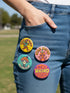 Set of 4 bright retro pinback buttons 2.25 inch. 1: Yellow button with cartoon Bigfoot in blue Hawaiian shirt and sunglasses striding under "Stay Weird" text. 2: Pink button with orange three-eyed alien in party hat holding a balloon. 3: Teal button with smiling mushroom in top hat riding a unicycle with "Stay Weird" banner. 4: Purple button with orange and yellow psychedelic 70s font reading "Stay Weird Forever." High gloss finish metal pins for backpacks, jackets, and weirdcore gifts.