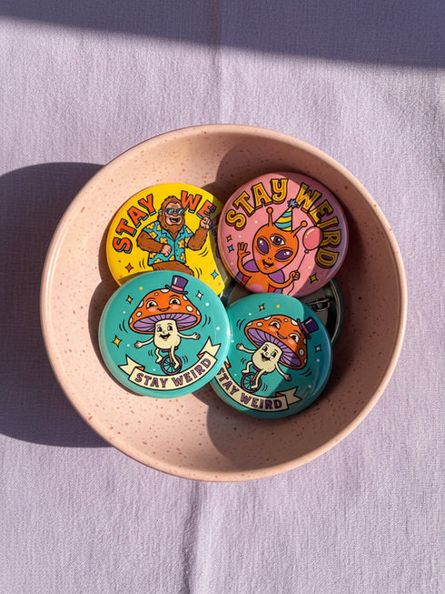Set of 4 bright retro pinback buttons 2.25 inch. 1: Yellow button with cartoon Bigfoot in blue Hawaiian shirt and sunglasses striding under "Stay Weird" text. 2: Pink button with orange three-eyed alien in party hat holding a balloon. 3: Teal button with smiling mushroom in top hat riding a unicycle with "Stay Weird" banner. 4: Purple button with orange and yellow psychedelic 70s font reading "Stay Weird Forever." High gloss finish metal pins for backpacks, jackets, and weirdcore gifts.