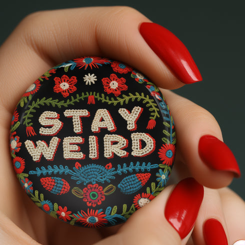 Stay Weird" black pinback button with pearl-effect lettering surrounded by folk art flowers in red and turquoise on a dark background.