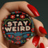 Stay Weird" black pinback button with pearl-effect lettering surrounded by folk art flowers in red and turquoise on a dark background.